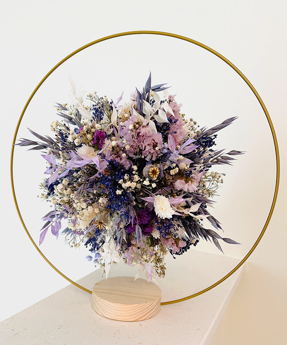 Trockenblumenball in lila Pastellfarben in einem goldfarbenen runden Metallrahmen auf einem hellen Holzsockel