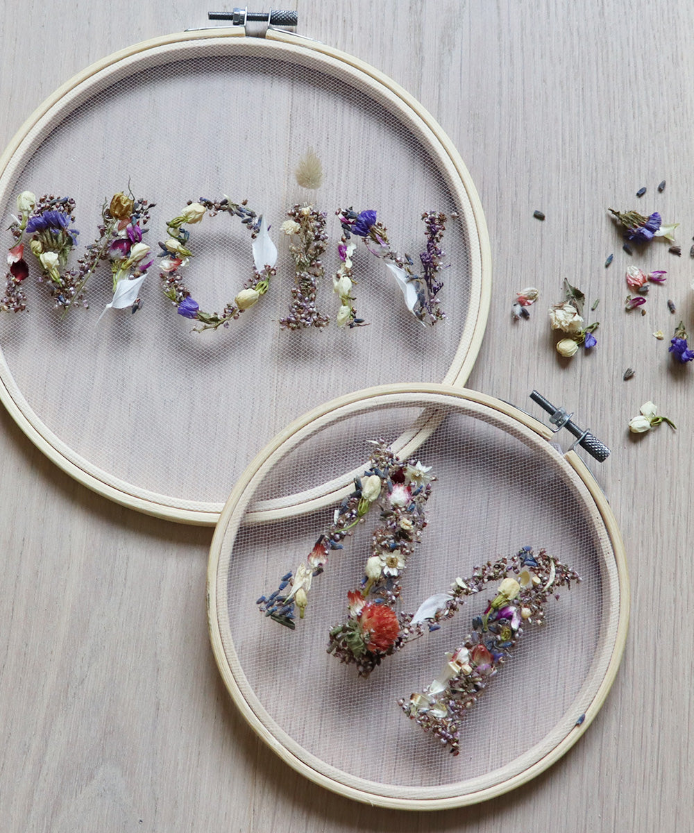 Zwei Stickrahmen mit getrockneten Blüten, die in einem Strickrahmen das Wort "Moin" und "M" formen