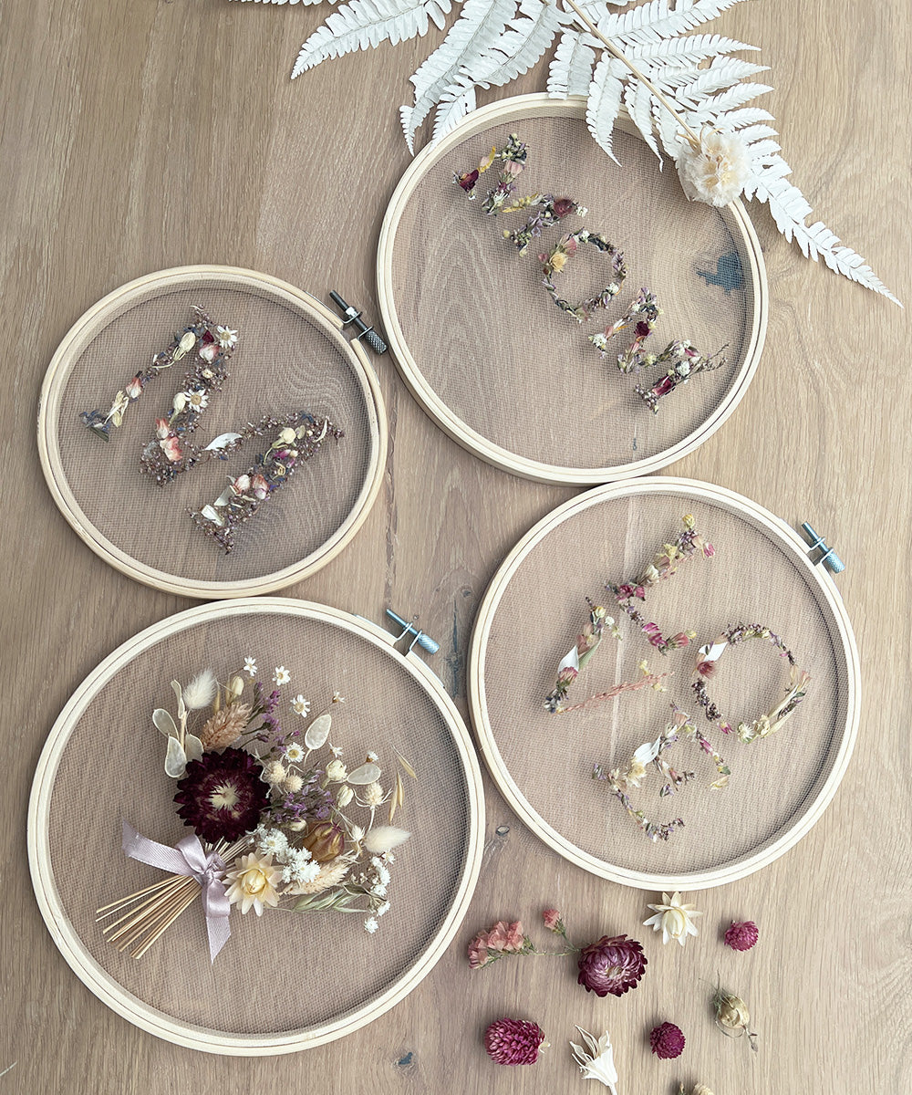 Vier Stickrahmen mit getrockneten Blumenarrangements auf hellem Holzuntergrund