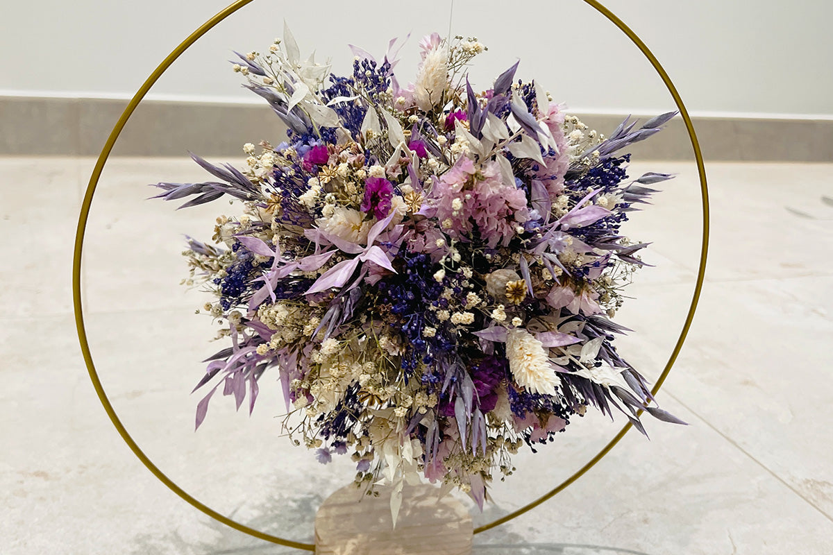 Trockenblumenball in lila Pastellfarben in einem goldfarbenen runden Metallrahmen auf einem hellen Holzsockel