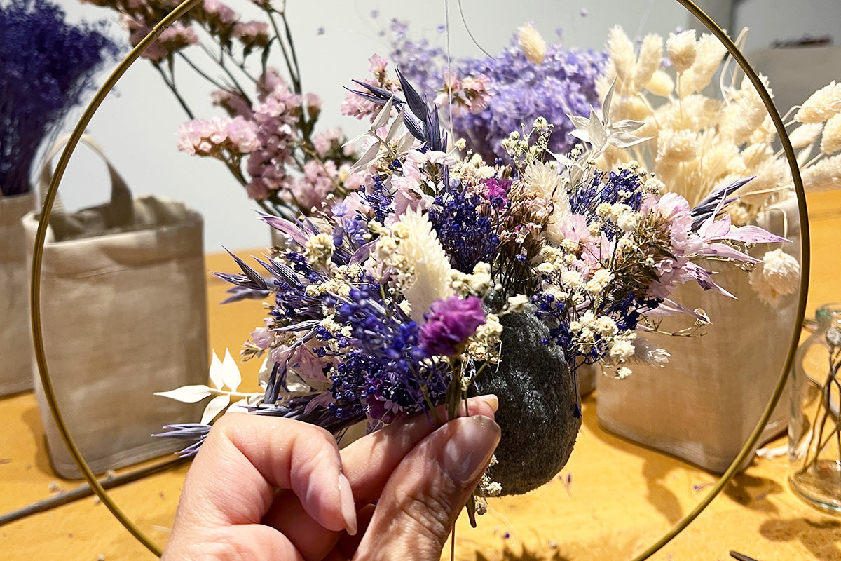 Trockenblumenball in lila Pastellfarben in einem goldfarbenen runden Metallrahmen und eine Hand mit Trockenblumen